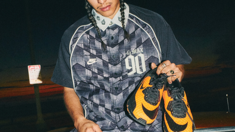 A man with braided hair wearing a patterned Nike Air Max 90 button-down shirt holding a pair of vibrant orange and black H...