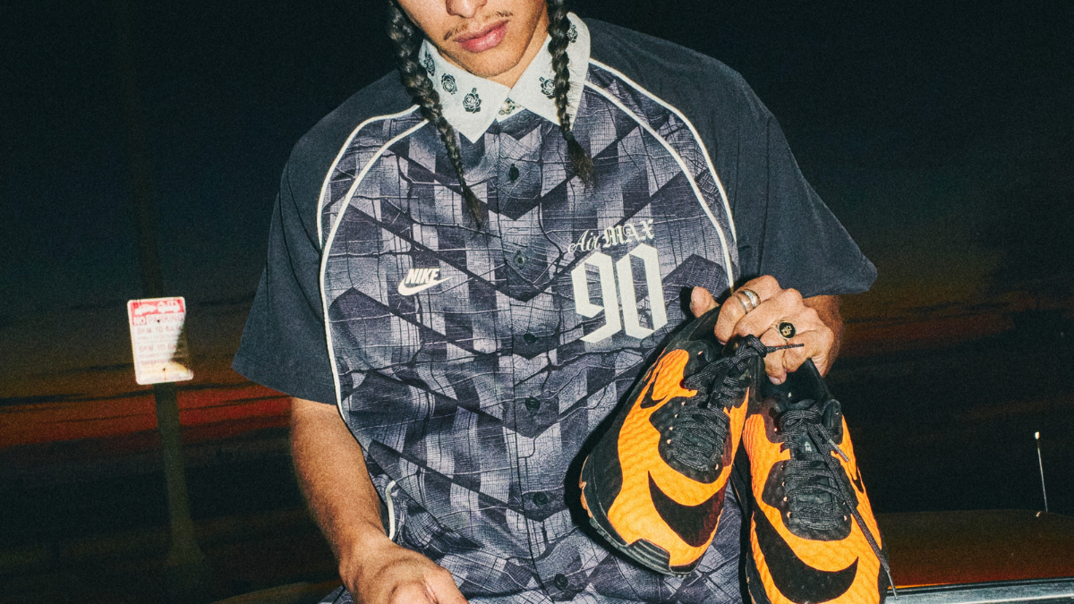A man with braided hair wearing a patterned Nike Air Max 90 button-down shirt holding a pair of vibrant orange and black H...