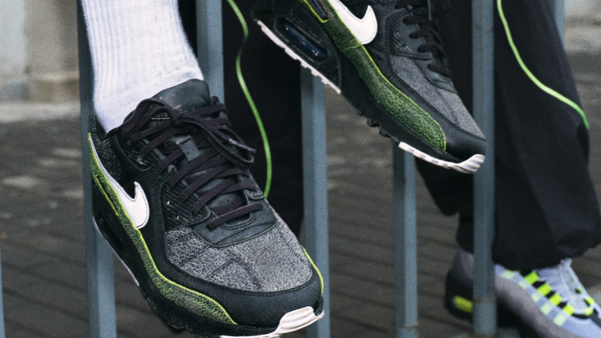 A close-up shot of feet resting on a metal fence wearing black Air Max 90 Mercurial sneakers featuring volt green accents and a classic football boot tongue.