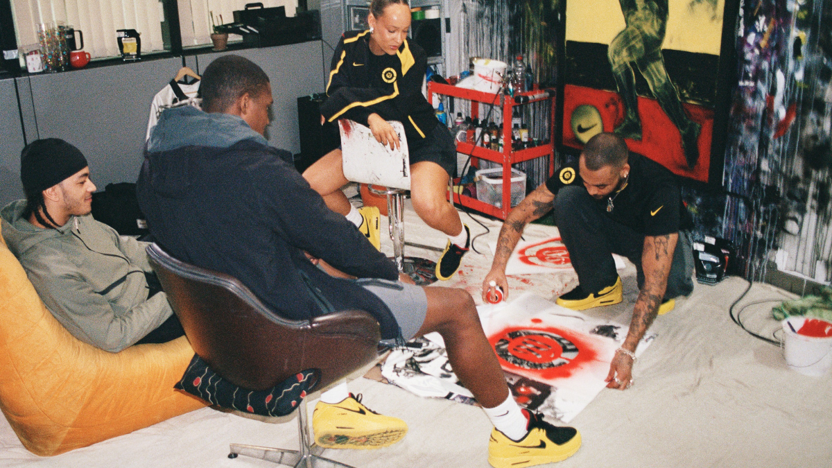 A diverse group of young creatives in a studio workshop setting, spray-painting stencils while wearing yellow and black Tiempo-inspired Air Max 90 sneakers.