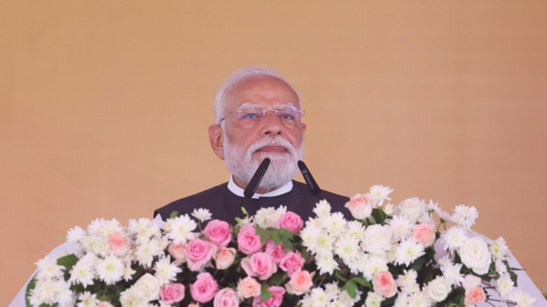 Prime Minister Narendra Modi speaking behind a podium covered in pink and white flowers during the Ganga Expressway launch.