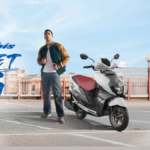 A young man standing next to a white and grey Suzuki Avenis scooter on a sunny urban rooftop in India.