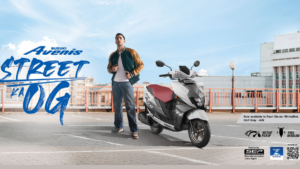 A young man standing next to a white and grey Suzuki Avenis scooter on a sunny urban rooftop in India.