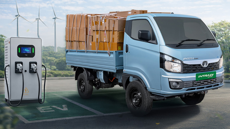 A light blue Tata Intra EV Pickup truck loaded with cargo boxes, connected to an EV charging station in an outdoor setting with wind turbines.