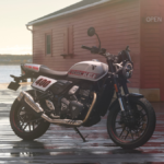 Side profile of the new Triumph Tracker 400 in White and Red with flat-track styling parked in front of a coastal shack.