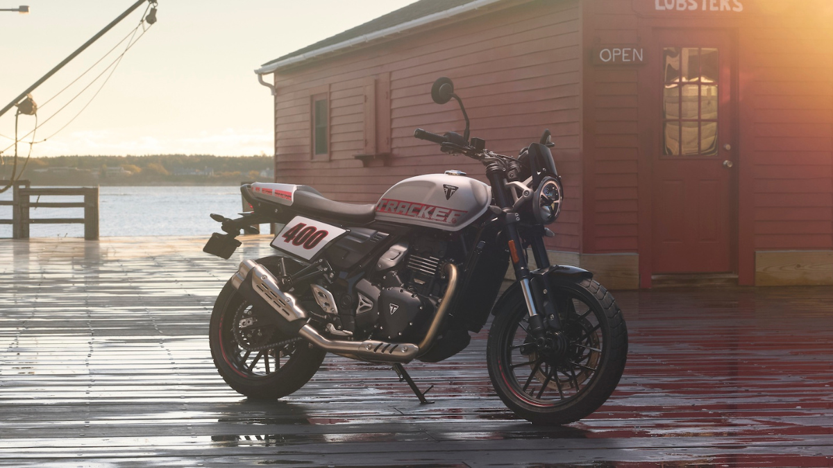 Side profile of the new Triumph Tracker 400 in White and Red with flat-track styling parked in front of a coastal shack.