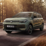 Front three-quarter view of the sage green Volkswagen Taigun driving on a sun-drenched forest road.