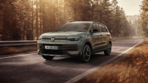 Front three-quarter view of the sage green Volkswagen Taigun driving on a sun-drenched forest road.