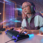 A woman with pink hair, glasses, and headset types on a Logitech G512 X keyboard with purple keycaps and a lit wrist rest,...