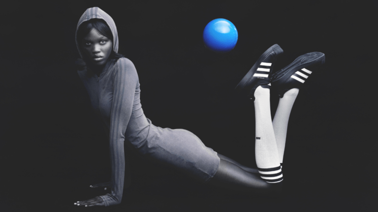 A model wearing a hooded dress, long white socks, and the black Modified Superstar shoes poses with a floating blue ball.