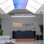 Modern office reception desk with a backlit buzzworks sign, white sofa, and indoor plants at the new Mysuru workspace.