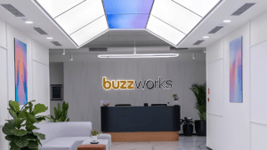 Modern office reception desk with a backlit buzzworks sign, white sofa, and indoor plants at the new Mysuru workspace.