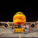 Night shot of a yellow and red DHL cargo jet head-on with a yellow tow tug on an airport ramp with crew. AI logistics.