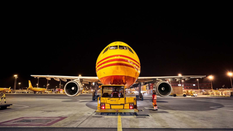 Night shot of a yellow and red DHL cargo jet head-on with a yellow tow tug on an airport ramp with crew. AI logistics.