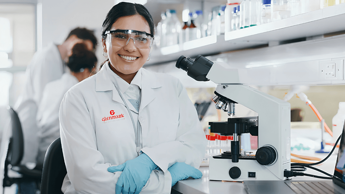 Smiling scientist in a Glenmark lab coat and safety glasses next to a microscope, representing the GLIPIQ launch in India.