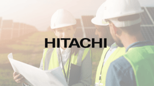 Engineers in safety gear review blueprints in a solar field with a large Hitachi logo overlaid, representing infrastructure.
