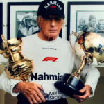 Jackie Stewart with cap and sweatshirt holding two large golden trophies; racing context.