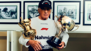Jackie Stewart with cap and sweatshirt holding two large golden trophies; racing context.