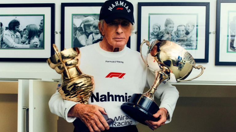 Jackie Stewart with cap and sweatshirt holding two large golden trophies; racing context.