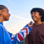 A person holding a small black JBL clip-on microphone towards a laughing friend outdoors for a mobile podcast.