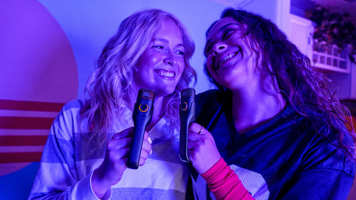 Two people singing into black JBL microphones held by ring handles under purple party lighting for karaoke.