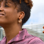 A woman wearing orange stem earbuds next to a floating touch-screen charging case for JBL audio context.