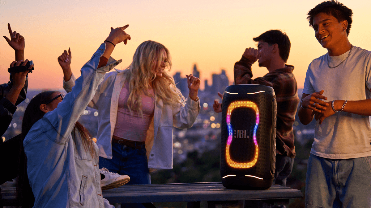 The pro-grade speaker built to last longer than your car 2 JBL PartyBox 130 on a wooden table at sunset with friends dancing and a city skyline in the background