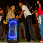 People dancing outdoors near a large JBL PartyBox 330 with blue neon lights during an evening party in India.