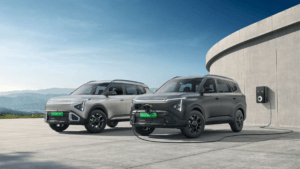 Silver and matte grey Kia Clavis EV SUVs parked side-by-side, the grey one connected to a wall charger in India.