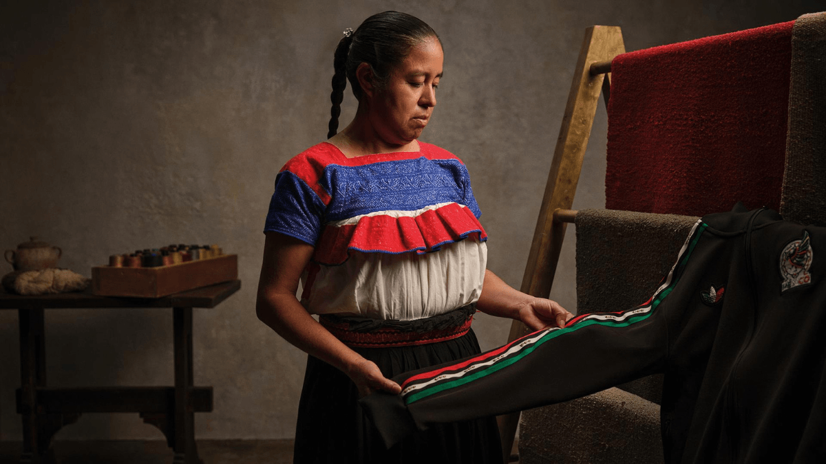 A female artisan in traditional clothing inspecting a black adidas track jacket with green, white, and red striped sleeve trim.