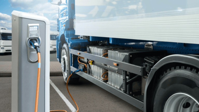 A blue heavy truck plugged into a silver charging station with an orange cable for Indian highway freight.