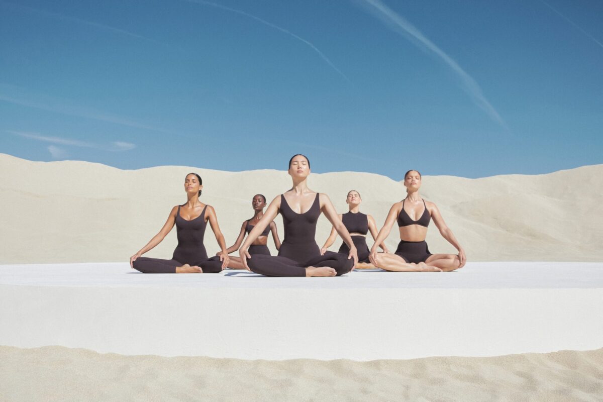 Nike and SKIMS introduce material that mimics a second skin 2 Five women of diverse ethnicities in matching dark brown NikeSKIMS workout gear sit in a row performing a meditative pose on a white platform.