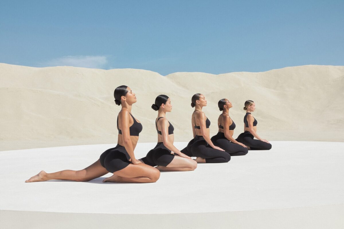 Nike and SKIMS introduce material that mimics a second skin 4 Five women in matching dark navy blue sports bras and high-waisted shorts perform a seated forward-facing pose with eyes closed on a white platform.