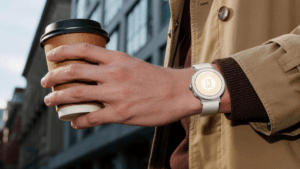 Realme Watch S5 in Sand White worn on a wrist holding a coffee cup outdoors, showing the round AMOLED screen display.