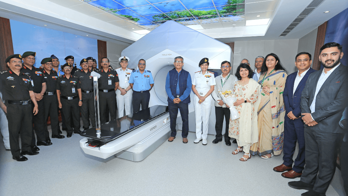 A large modern ring gantry medical machine surrounded by a group of military officials and civilians in a hospital room.