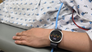 Samsung Galaxy Watch on a patient's wrist in a hospital gown, displaying a colourful health waveform and '59%'.