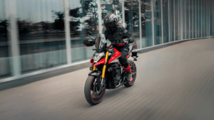 Rider in black gear on a red Ultraviolette electric crossover passing a modern glass building on an Indian city street.