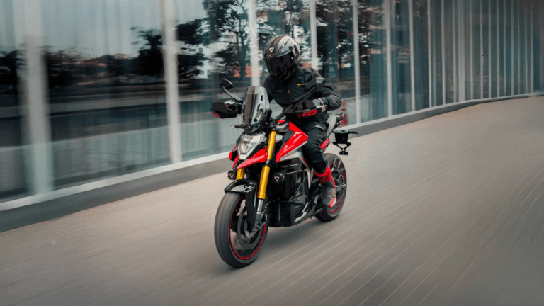 The electric motorcycle that justifies ditching petrol 2 Rider in black gear on a red Ultraviolette electric crossover passing a modern glass building on an Indian city street.