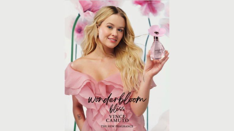 Woman in pink ruffled dress holds Vince Camuto Wonderbloom Bliss bottle with soft petal cap. Pink paper flowers are in bac...