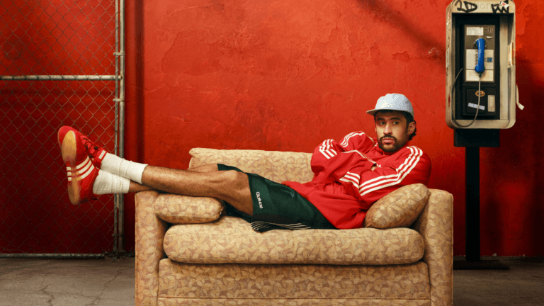 Bad Bunny lounging on an armchair wearing the red adidas Ballerina sneaker against a vivid red wall backdrop.