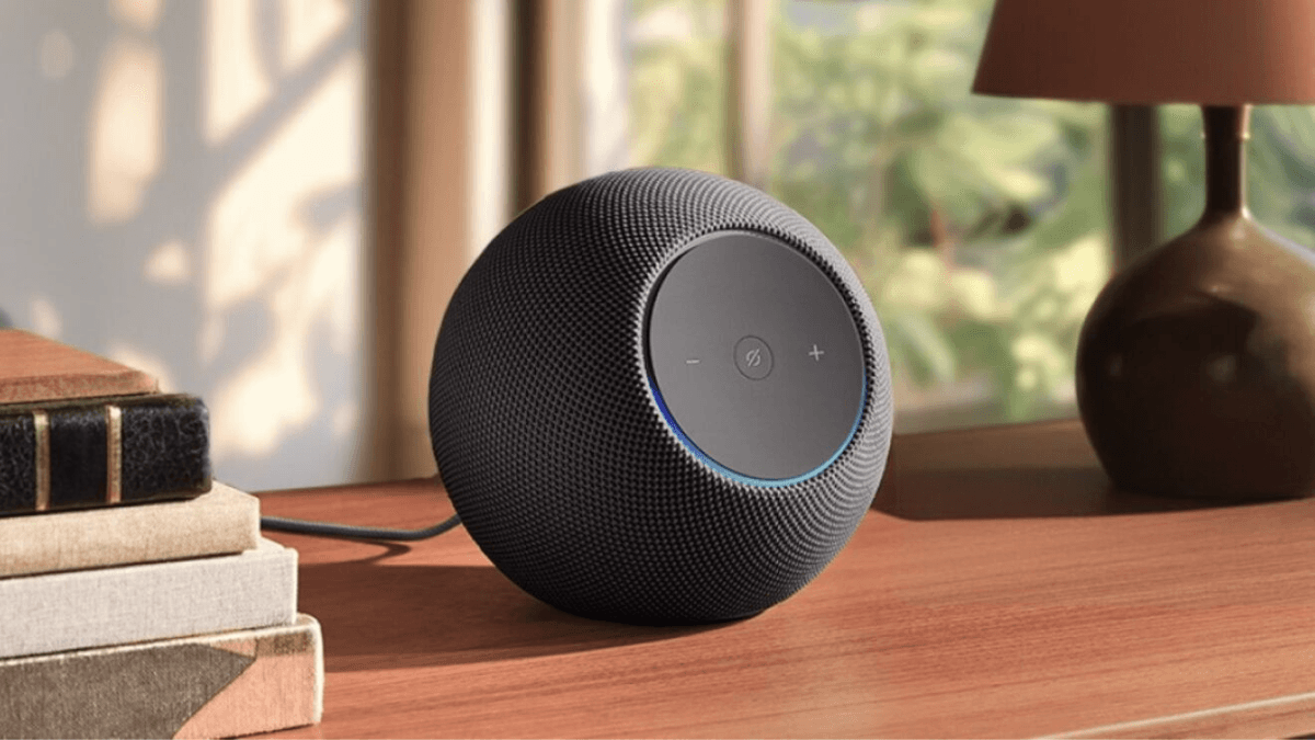 A grey spherical Amazon Echo smart speaker on a wooden desk next to a stack of books and a lamp in an Indian home.