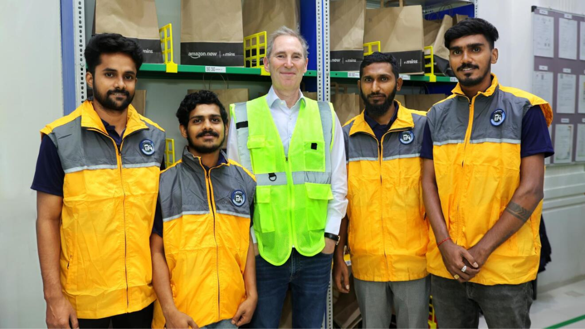 Delivery in minutes reaches 300 Indian cities this year 2 Four delivery associates in yellow vests and a man in a green vest stand before shelves holding Amazon Now bags in India.
