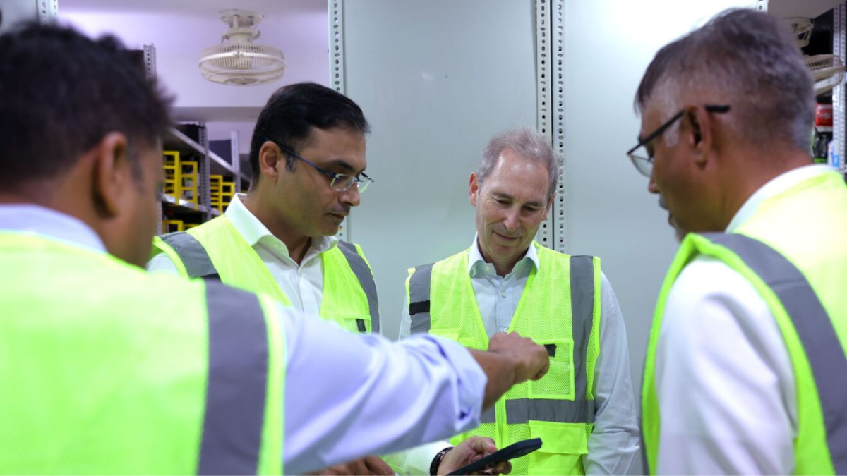 Delivery in minutes reaches 300 Indian cities this year 1 Four men in green safety vests look at a smartphone inside a warehouse storage area with metal shelving in India.