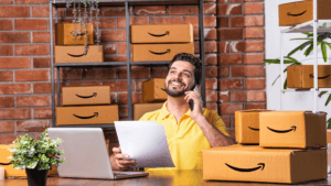 A smiling man in a yellow shirt talks on a phone at a desk with a laptop and Amazon boxes, managing his seller store.