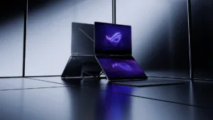 Two grey ASUS laptops on a dark reflective surface, one showing purple logos on dual OLED screens.