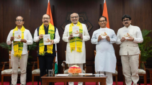 Five men stand holding books in front of a wooden panel and flags, three wearing yellow patterned scarves.