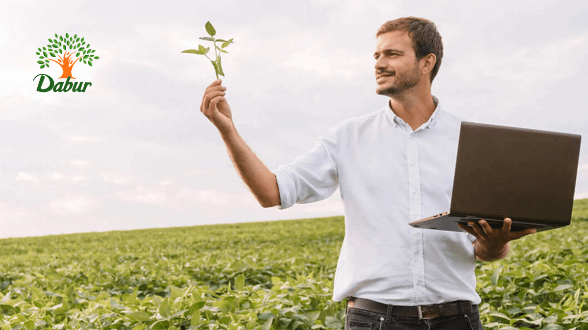 With four ₹1,000 crore brands this FMCG giant is betting on AI 1 A man in a field holding a plant stem and a laptop with a Dabur logo, representing digital modernisation.