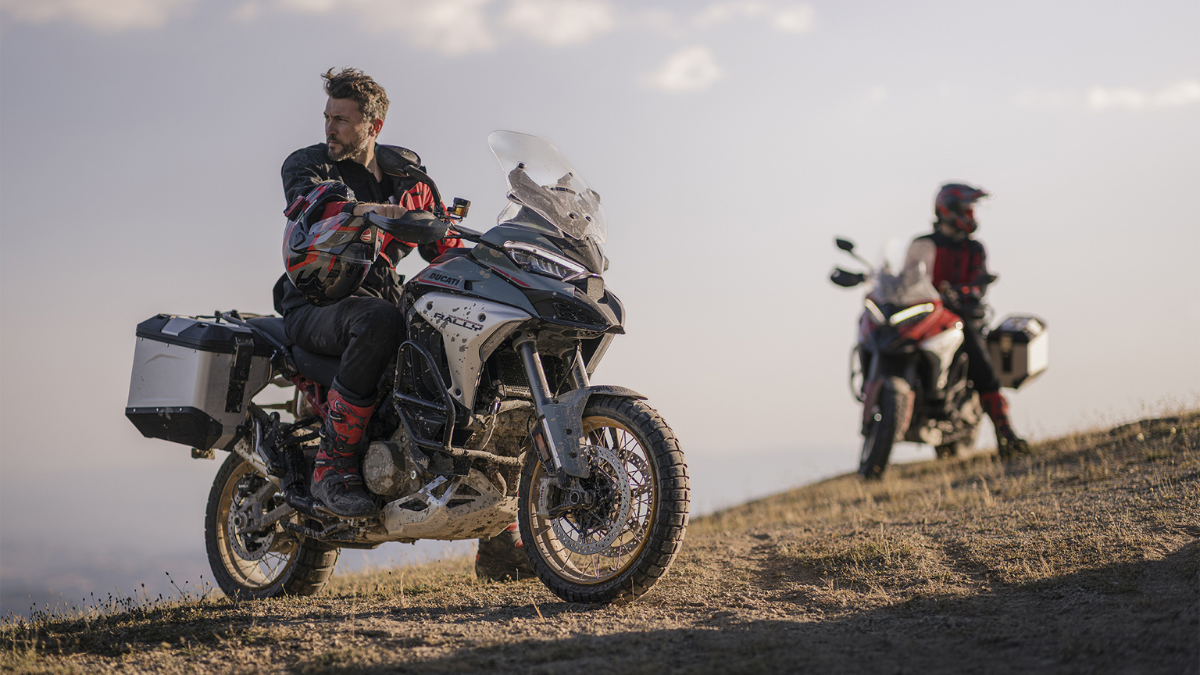 Rider on a green motorcycle with side cases on a dirt path with a second rider on a red motorcycle behind.