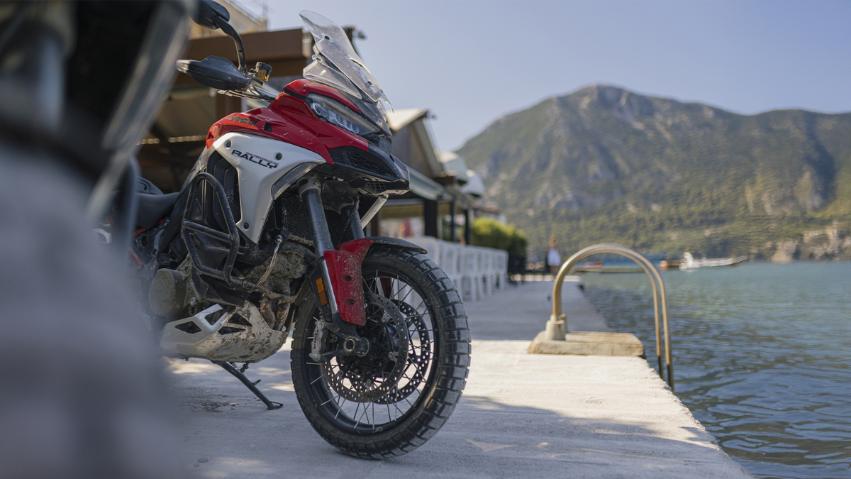 Blurred foreground object next to a red adventure motorcycle parked on a concrete pier by a lake and mountains.