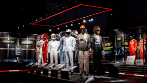 Race suits worn by famous drivers displayed on mannequins inside a dark museum gallery next to a red racing car.
